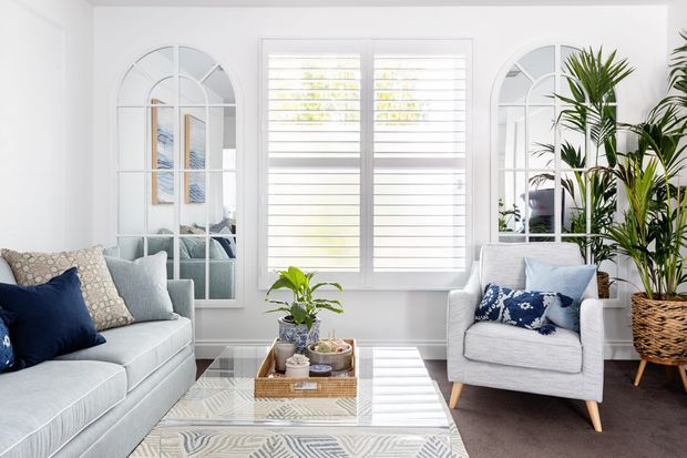 Bright, refined living room with a pale upholstered sofa and armchair, patterned blue and neutral cushions, a glass-topped coffee table, indoor plants, arched mirrored wall panels, and white plantation shutters. The room has a light, airy palette with dark carpet providing contrast.