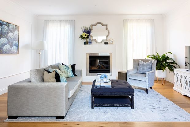 Bright, refined living room with a neutral upholstered sectional sofa, accent armchair, tufted ottoman, pale patterned area rug, white walls with panel moulding, and a central fireplace with a decorative mirror above it. Tall sheer-curtained windows provide abundant natural light, while light timber flooring and restrained blue-grey accents create an airy, elegant atmosphere.