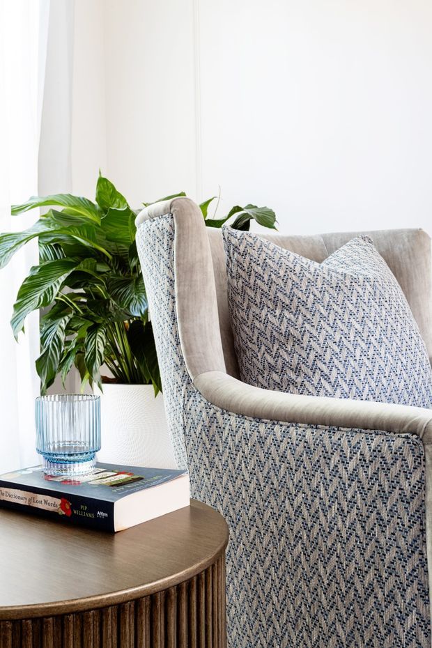 Close-up interior vignette featuring a textured upholstered armchair in cream and blue geometric fabric beside a round fluted timber side table. A leafy indoor plant in a ribbed glass vessel, books, and bright white walls create a fresh, refined seating corner.