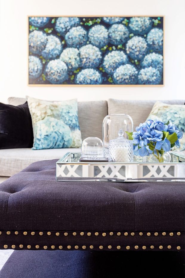 A styled living room vignette featuring a pale upholstered sofa with blue floral cushions, a large framed blue floral artwork, and a deep purple tufted ottoman with brass nailhead trim. A mirrored tray holding glass cloches, crystal décor, and blue hydrangeas sits on the ottoman. The palette is bright and refined, with soft neutrals, blue accents, and dark plum upholstery.