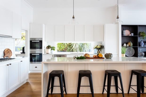 Bright, spacious white kitchen with shaker-style cabinetry, a large pale stone island with seating for four, integrated appliances, pendant lights, timber flooring, and a dark open display cabinet. Natural greenery is visible through the windows, while decorative objects and produce add warmth to the otherwise clean scheme.
