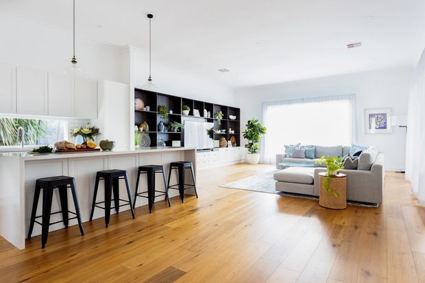 Bright open-plan kitchen and living room with white cabinetry, a long island with black bar stools, black built-in shelving, timber flooring, and a grey sectional sofa. Large windows provide abundant natural light, creating an airy contemporary interior.