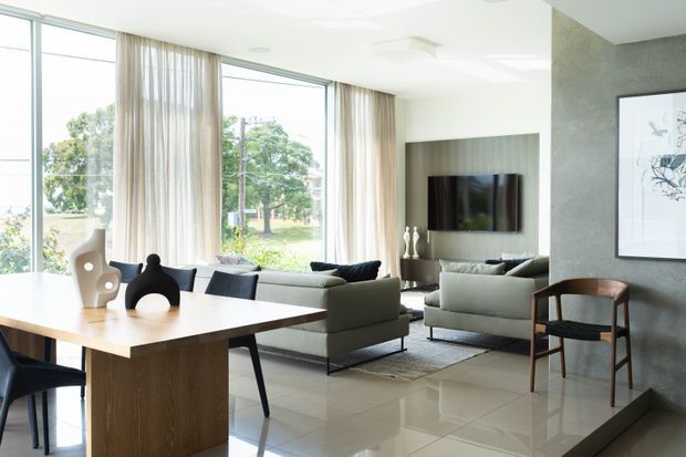 Bright open-plan living and dining area with floor-to-ceiling glazing, sheer curtains, a timber dining table, contemporary upholstered seating, tiled flooring, and a wall-mounted television set within a textured feature wall. The room has a calm, refined, neutral aesthetic with abundant natural light.