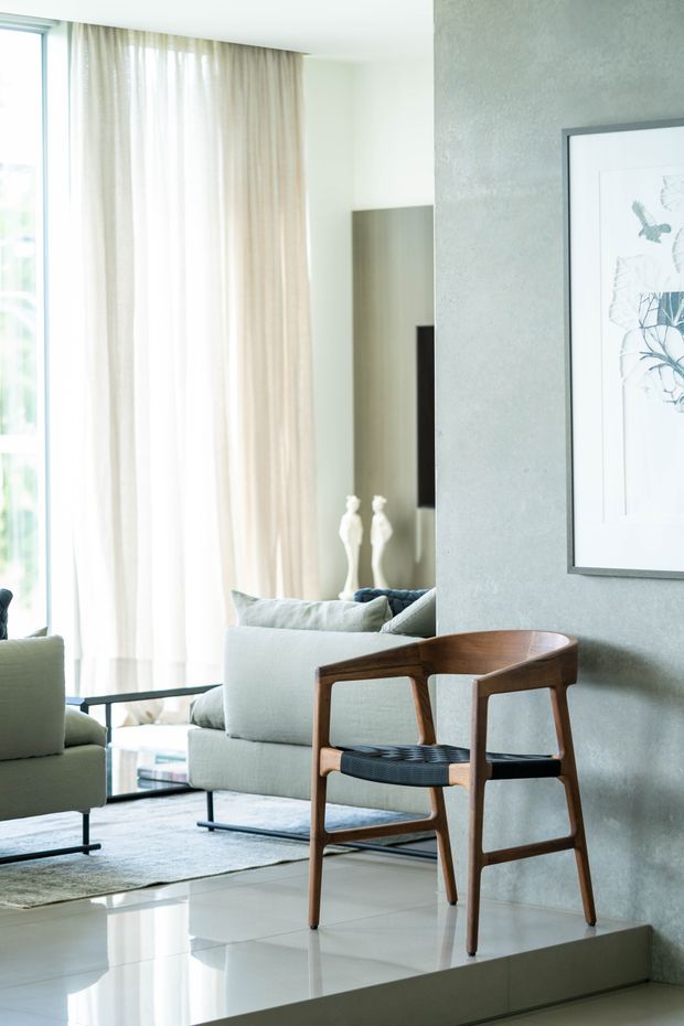 Bright, refined living room with floor-to-ceiling sheer curtains, pale upholstered seating, a textured grey feature wall, polished light flooring, and a sculptural mid-century-style wooden chair with woven seat. A framed botanical artwork and television are visible, with the living area raised slightly above the adjacent floor.