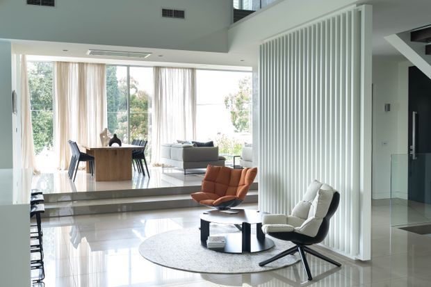 Bright, open-plan contemporary interior with a sunken living area, dining space, and expansive floor-to-ceiling glazing. A white vertical slatted partition separates the living zone from the circulation area, while polished light flooring, sculptural lounge chairs, a round rug, and a low coffee table create a refined mid-century-inspired seating arrangement.