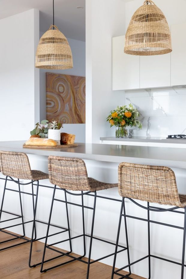 Bright contemporary kitchen with a white island and benchtop, integrated white cabinetry, pale marble-look splashback, timber flooring, and three woven rattan bar stools with black metal frames. Large woven pendant lights add a natural textural element, while a floral arrangement and artwork provide decorative accents.