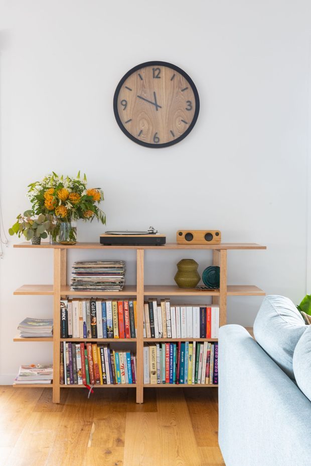 Bright living room corner featuring a light timber open bookshelf filled with books and magazines, a turntable and compact speaker on top, decorative vessels, a vase of flowers, and a pale blue sofa. A large round wood-faced wall clock hangs above the shelving against a white wall, with warm timber flooring below.