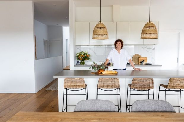 Bright contemporary open-plan kitchen viewed from the dining area, featuring a large pale benchtop island with four woven bar stools, white flat-panel cabinetry, a white tiled splashback, timber flooring, and two woven pendant lights. A person stands behind the island, with a hallway visible to the left.