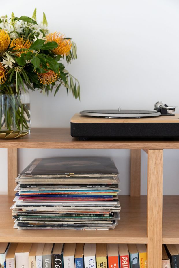Close-up interior detail of a natural timber open shelving unit used as a media console and bookcase. A turntable sits on the upper shelf beside a large floral arrangement, with stacked vinyl records and books stored below. The scene has a warm, curated, music-focused character against a plain light wall.