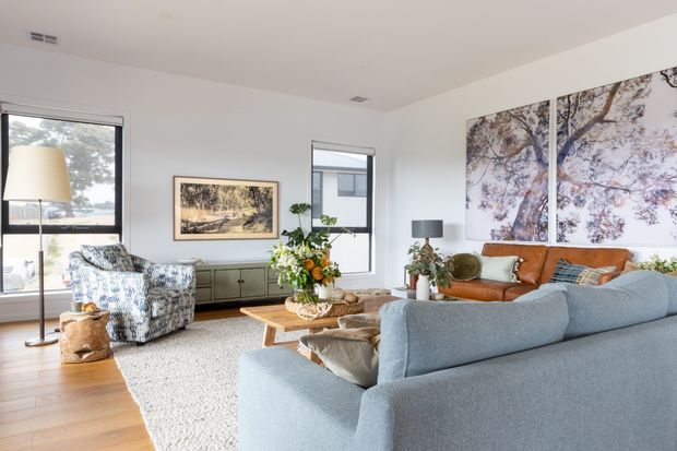 Bright, spacious contemporary living room with white walls, warm timber flooring, a large textured rug, a pale blue-grey sectional sofa, brown leather sofa, patterned armchair, timber coffee table, indoor plants, and layered framed artwork. Large windows provide abundant natural light, creating an airy and inviting atmosphere.
