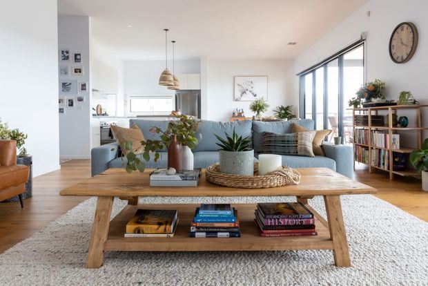 A bright, open-plan living room with a large pale blue sectional sofa, a substantial rustic timber coffee table, woven rug, indoor plants, bookshelves, and expansive black-framed windows. A compact kitchen and dining area are visible beyond the seating zone. The room has light timber flooring, white walls, soft neutral furnishings, and contemporary coastal styling.