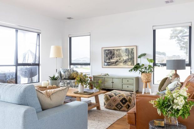 Bright, airy living room with large windows, pale upholstered seating, a cognac leather armchair, timber coffee table, patterned rug and ottoman, indoor plants, and a low mid-century-style sideboard. The room has white walls and ceiling, timber flooring, and abundant natural light.