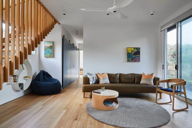 Bright contemporary living room with warm timber flooring, a large brown leather sofa, round coffee tables, a sculptural grey rug, and a timber slatted stair balustrade. Full-height glazing opens to a leafy exterior, while artwork and a blue lounge chair add colour.
