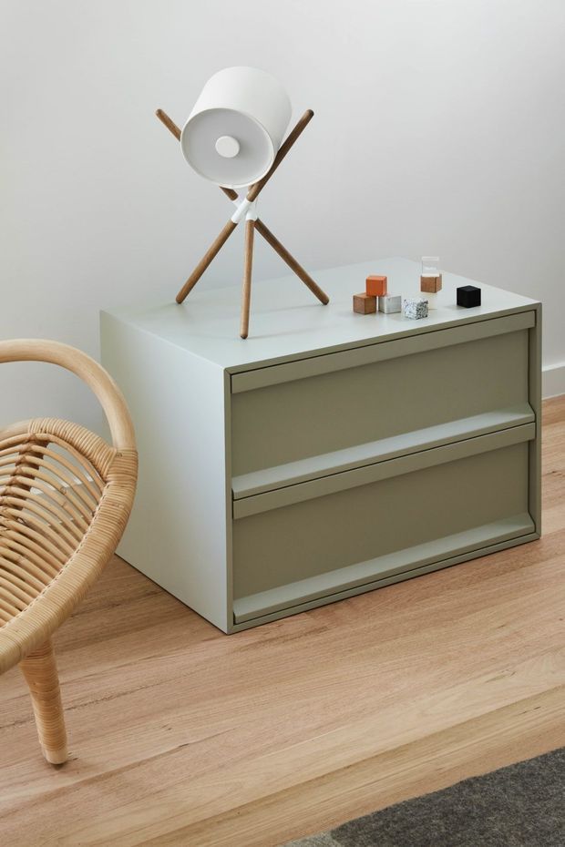 A minimalist interior vignette featuring a muted olive-grey two-drawer cabinet on a light timber floor. A white mid-century tripod table lamp with wooden legs sits on top beside small decorative cubes, while part of a woven rattan chair is visible at the left edge. The scene is softly lit with a restrained neutral palette and clean, simple forms.