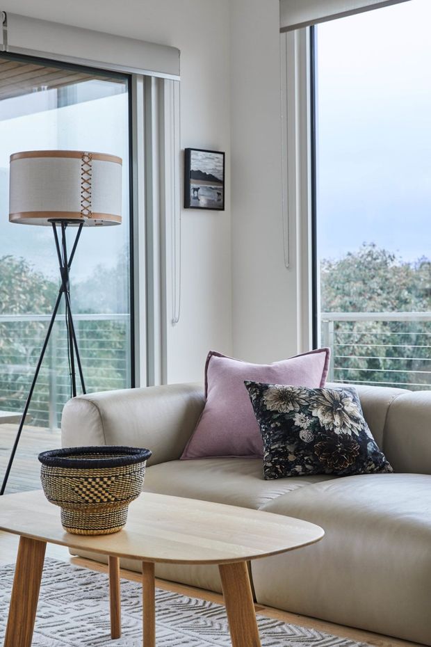 Bright, softly styled living room with a cream upholstered sofa, blush and floral cushions, a pale timber coffee table, patterned rug, woven basket, and a slender tripod floor lamp. Full-height glazed doors and windows open onto a balcony with cable railing and leafy views, creating an airy contemporary setting.