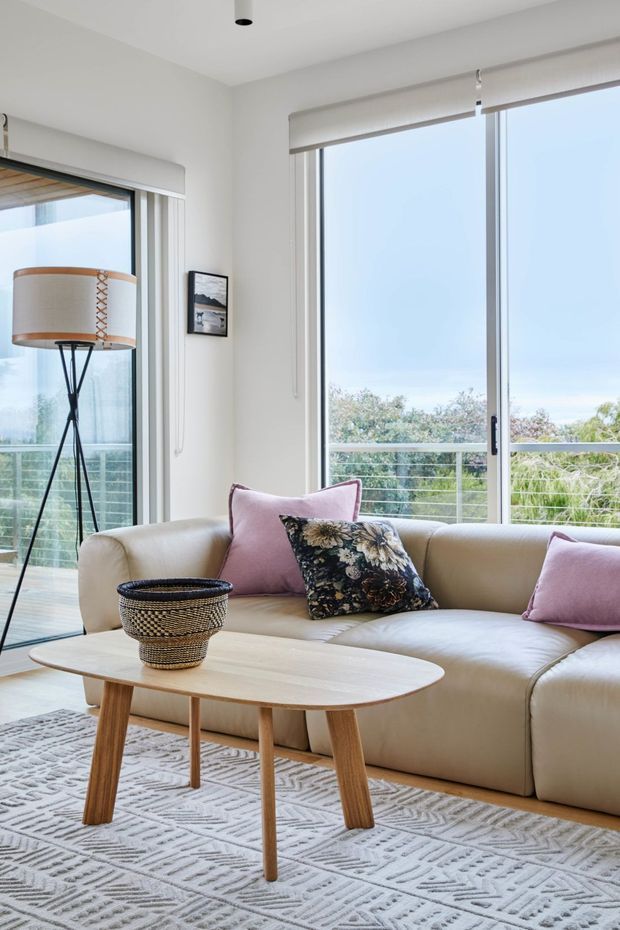 Bright, airy living room with a cream sectional sofa, pale timber coffee table, patterned light rug, woven basket, tripod floor lamp, and blush and floral cushions. Full-height glazed doors and windows open to a leafy exterior balcony or terrace, with roller blinds and slim metal frames. The room has a clean contemporary design with soft neutral tones and natural textures.