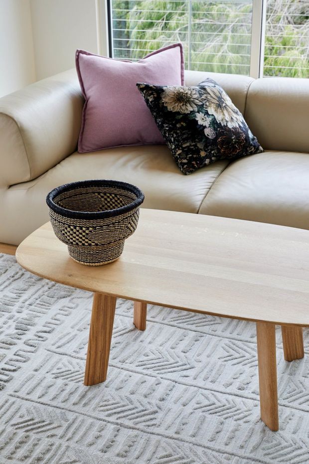 Bright living room scene featuring a cream leather sofa with mauve and patterned floral cushions, a pale timber oval coffee table, a black-and-natural woven decorative basket, and a light textured geometric rug. Large windows with horizontal security bars overlook leafy greenery.