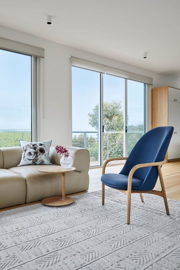 Bright contemporary living room with expansive floor-to-ceiling glazing, pale timber flooring, a cream upholstered sofa, a sculptural blue lounge chair, and a large patterned neutral rug. Roller blinds, white walls, recessed ceiling spotlights, and built-in timber-and-white cabinetry create a clean, airy interior overlooking greenery and water.