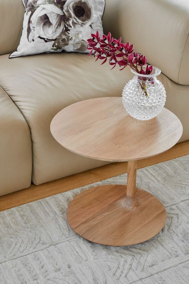 Close-up interior vignette featuring a light beige leather sectional sofa, a round light timber pedestal side table, a textured clear glass vase with burgundy foliage, and a pale patterned area rug. The styling is neutral, warm, and refined.
