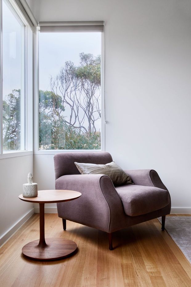 A bright, compact sitting area with a sculptural upholstered armchair beside a small round timber side table. Large corner windows provide abundant natural light and overlook trees. The room has plain white walls, light timber flooring, and a restrained neutral palette.