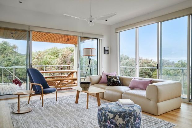 Bright, airy living room with extensive floor-to-ceiling glazing opening onto a timber balcony or deck. The room features a cream sectional sofa, blue upholstered armchair, light timber coffee tables, patterned ottoman, large area rug, floor lamp, and a white ceiling fan. Natural vegetation and distant landscape are visible beyond the glazing.
