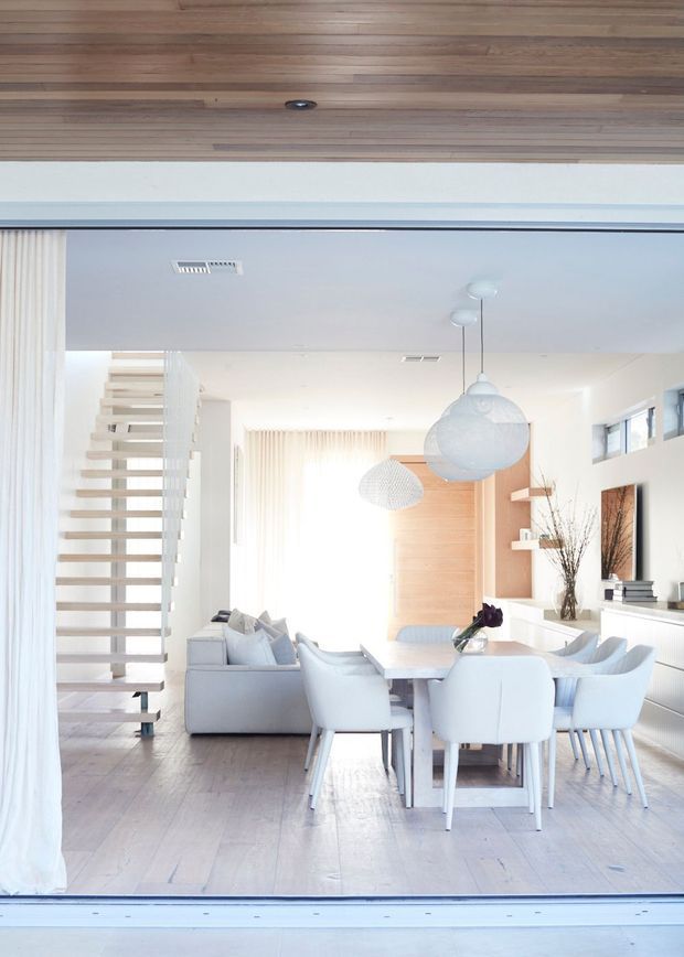 Bright open-plan interior viewed through a large glazed opening, combining a dining area and living room. The space features pale timber flooring, white walls and joinery, a sculptural open-riser staircase, soft upholstered seating, sheer curtains, and multiple pendant lights. Warm timber ceiling lining and a timber feature wall add natural contrast to the predominantly white scheme.