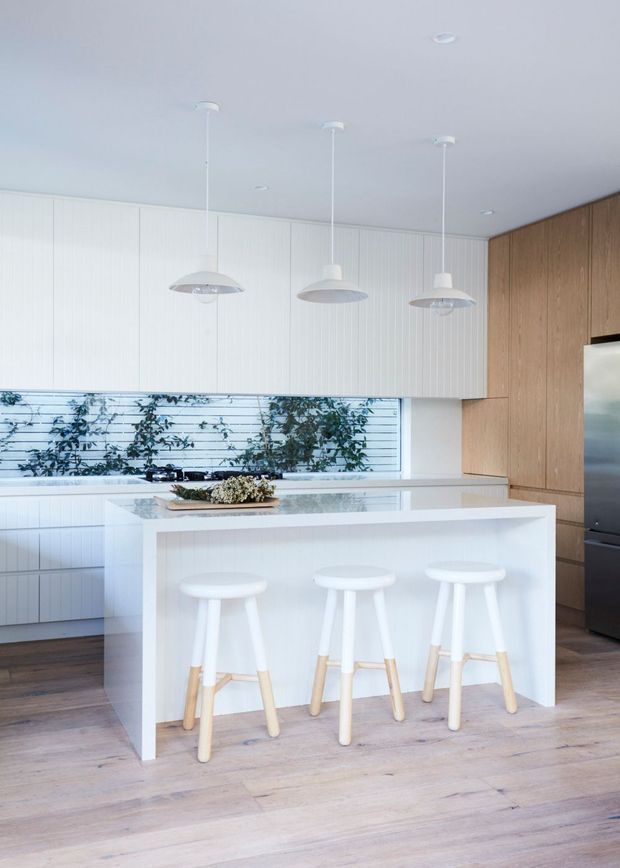 Bright contemporary kitchen with a large white island and three stools, white vertically panelled cabinetry, warm timber cabinetry on the right wall, pale timber flooring, integrated appliances, and three pendant lights. A horizontal window above the benchtop provides views of greenery outside.