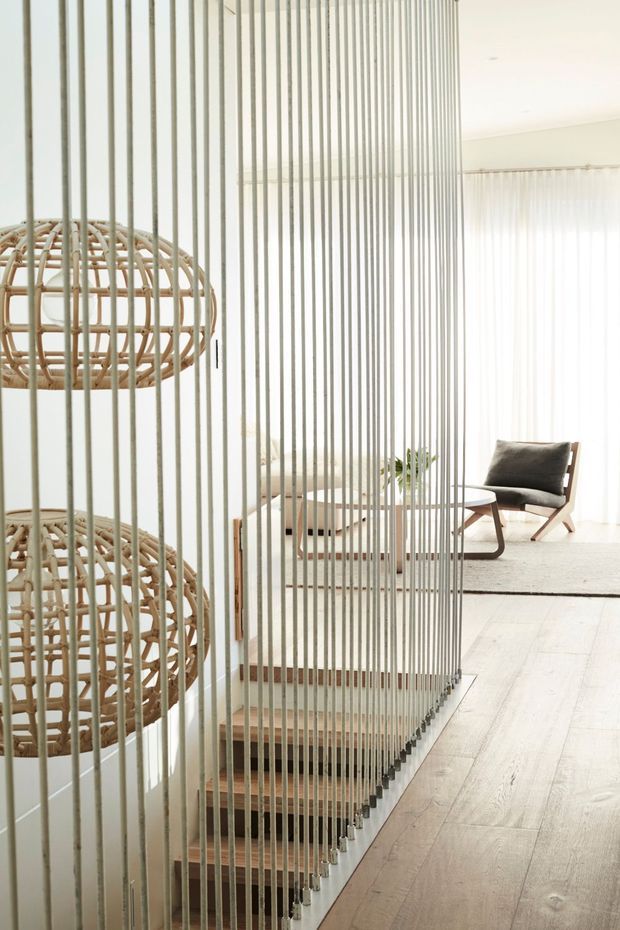 Bright, refined contemporary interior showing a staircase screened by closely spaced vertical rods, with timber stair treads and pale timber flooring. Woven rattan pendant lights hang beside the stair, while a lounge area with a dark upholstered armchair, small round table and sheer curtains is visible beyond. The palette is predominantly white, cream and light wood with natural woven accents.