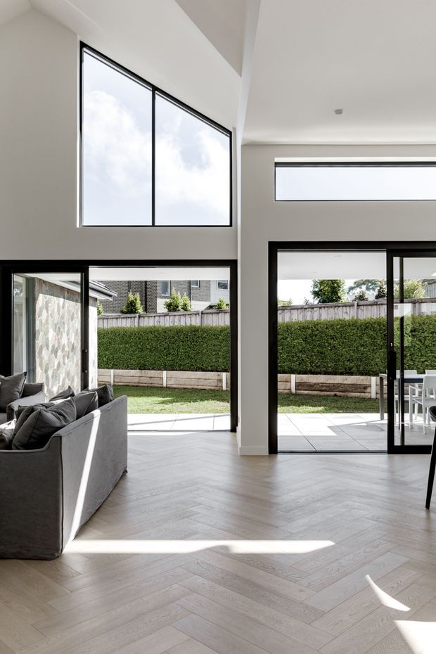 Bright contemporary living room with a high ceiling and extensive black-framed glazing opening onto a tiled patio and landscaped backyard. A grey sectional sofa occupies the left side, while the pale timber-look herringbone floor and white walls create a light, airy interior. Clerestory windows and large upper windows bring in abundant daylight.