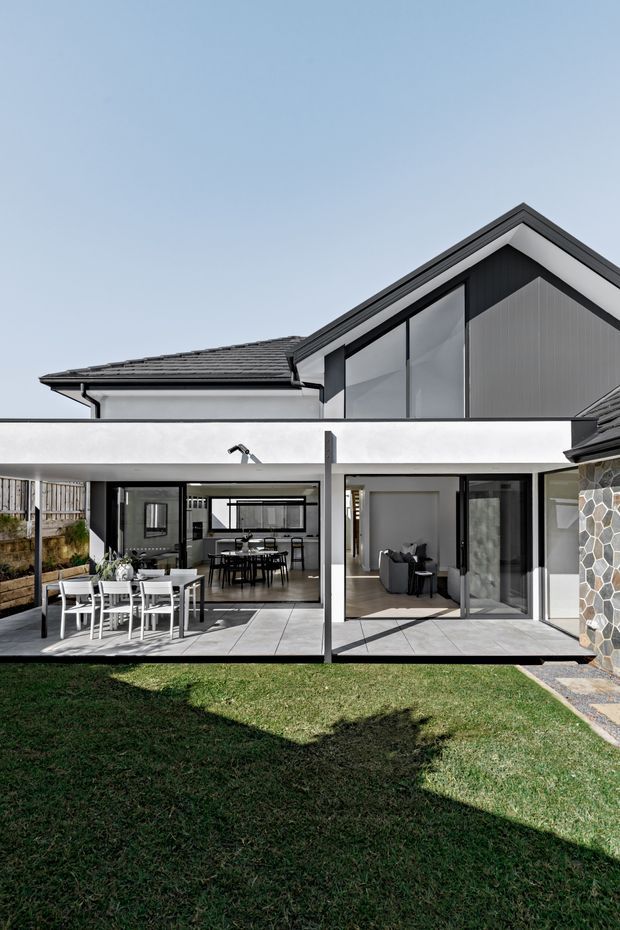 A modern two-storey residential house viewed from the rear courtyard. The exterior combines dark pitched roofs, white rendered walls, black-framed glazing, and vertical dark cladding. Large sliding glass doors open onto a light tiled terrace with outdoor dining furniture, while the interior dining and living spaces are visible through the glazing. A stone-clad wall frames the right side of the image, with a lawn in the foreground.