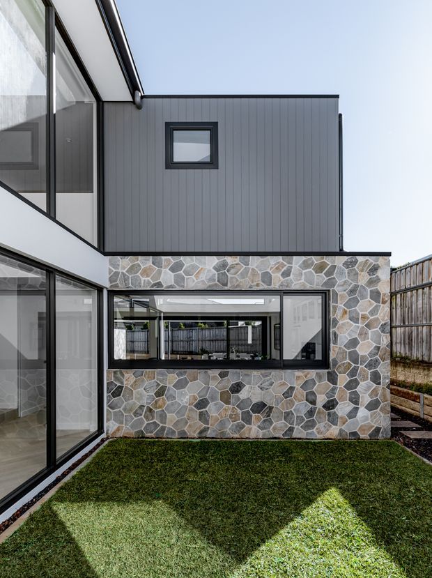 A contemporary multi-level residential courtyard viewed from outdoors, with dark vertical cladding, extensive black-framed glazing, and a prominent wall finished in irregular natural stone. A small lawn occupies the foreground, with timber fencing along the side boundary. The composition highlights the building facade, courtyard-facing windows, and contrasting geometric materials.