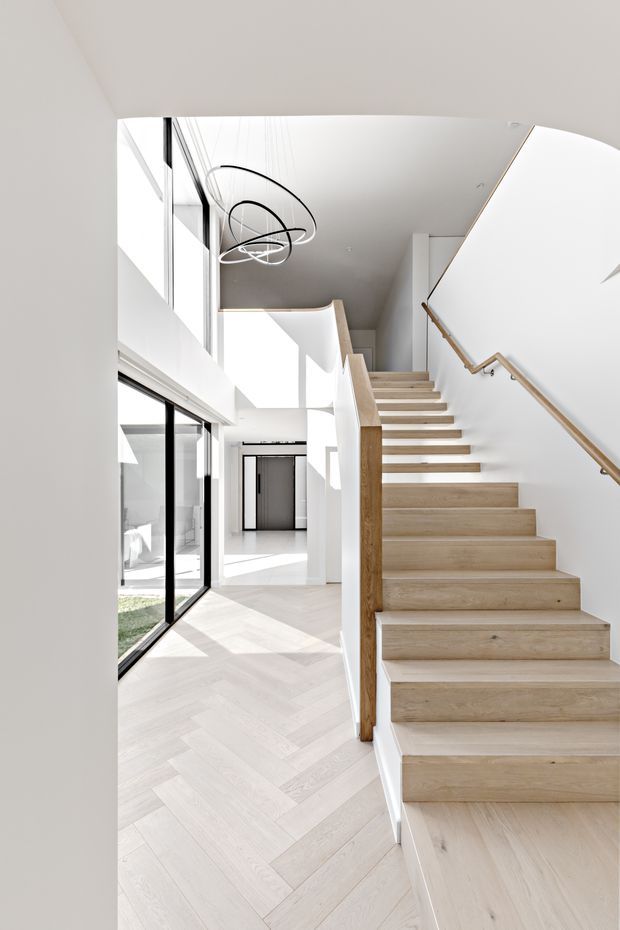 Bright, minimalist residential stairway viewed from an open foyer or hallway. A broad timber staircase with a simple timber handrail rises beside a white balustrade, while pale herringbone timber flooring extends through the lower level. Full-height black-framed glazing brings in strong natural light, and a sculptural loop pendant hangs over the stair void. White walls, high ceilings, clean lines, and layered open circulation create an airy contemporary interior.
