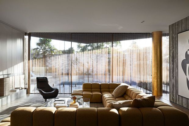 A spacious, sunlit contemporary living room with a large modular brown sectional sofa arranged around a low coffee table, a sculptural lounge chair, and expansive floor-to-ceiling glazing. Timber screening and outdoor greenery are visible beyond the glass, while a cylindrical timber-clad column and dark textured wall finish add architectural interest.
