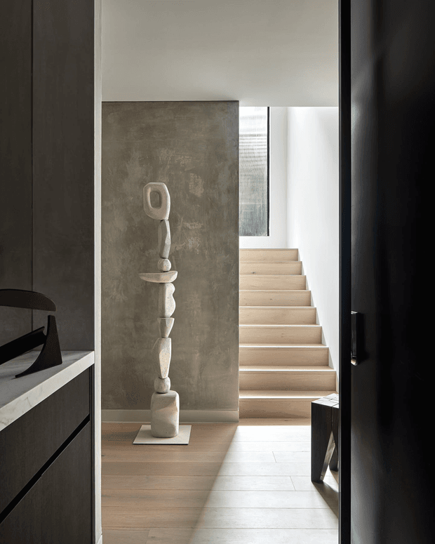 A refined contemporary interior view from a dark joinery-lined passage toward a light timber staircase. The space features pale timber flooring and stair treads, a textured grey feature wall, strong black vertical door or wall framing, and a tall abstract stone sculpture displayed against the wall. Natural light enters from the stairwell and upper glazing, creating a calm, gallery-like atmosphere.