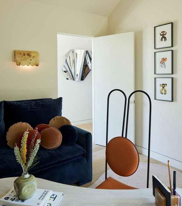 A warmly lit, artfully styled living room with a deep navy velvet sofa, rounded rust and ochre cushions, a pale timber coffee table, and a sculptural black metal-and-rust leather lounge chair in the foreground. Cream walls and light timber flooring create a soft neutral backdrop, with framed artwork, a wall sconce, and an open white door adding visual interest.
