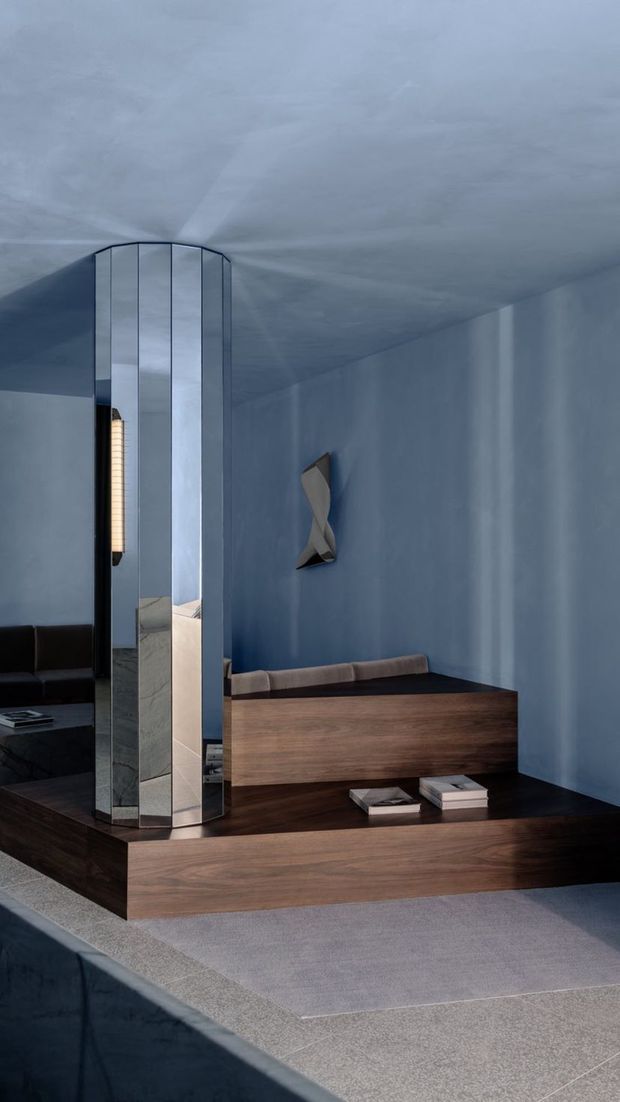 A moody, refined interior seating area with a dark timber platform and built-in timber bench against a blue-grey wall. A tall faceted mirrored architectural screen or column separates the seating from an adjacent dark sofa area, while sculptural wall lighting and books add understated detail. The space uses polished reflective surfaces, dark wood, blue-grey plastered walls, and soft grey carpet.