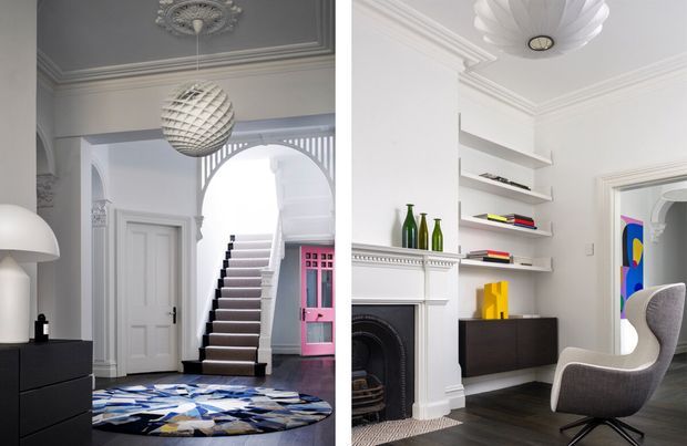 Split view of a refined, light-filled interior featuring a traditional hallway and staircase on the left, and a contemporary living or study area on the right. The spaces have white decorative mouldings, ceiling roses, arch details, dark timber flooring, sculptural pendant lights, built-in shelving, a fireplace, and modern furnishings with colourful accents.