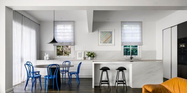 Bright contemporary open-plan kitchen and dining area with white cabinetry, a pale stone island and splashback, dark timber flooring, a black pendant light, and bold blue dining chairs. A mustard-yellow sofa is partially visible in the foreground, with sheer curtains and windows adding natural light.