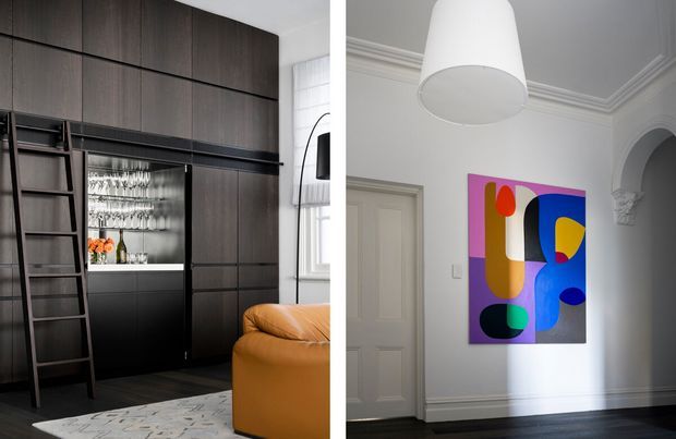 Split view of a refined interior featuring dark full-height built-in cabinetry with integrated shelving, ladder, display storage and glassware, paired with a bright room with ornate white cornices, panelled doors, dark timber flooring and a colourful abstract artwork. A tan leather sofa and large white pendant add warmth and contrast.