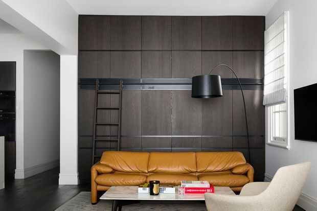 A compact contemporary living room with a warm tan leather sofa, sculptural floor lamp, cream lounge chair, low coffee table, dark timber wall panelling with integrated ladder shelving, dark flooring, and a wall-mounted television. White walls and a partially covered window provide contrast.