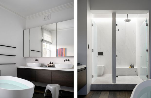 A contemporary bathroom with a double vanity, freestanding bathtub, and a spacious walk-in shower. The room combines white walls and sanitaryware with dark timber cabinetry and dark flooring, featuring frameless glass shower panels, a central partition, wall-mounted taps, and simple black hardware.