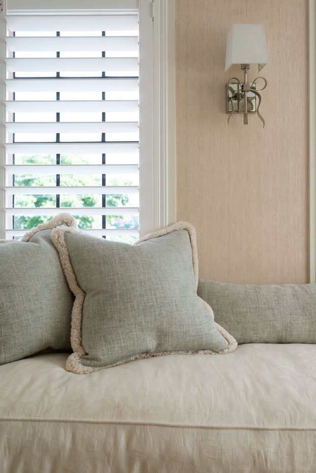 Close-up interior detail of a softly styled upholstered sofa or daybed beneath a window with white horizontal plantation shutters. Pale sage-grey cushions with textured cream edging sit against a light neutral wall, while an ornate metallic wall sconce adds a small decorative feature. The overall composition is calm, refined, and softly layered.