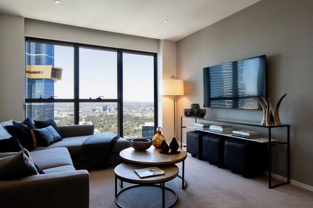 A refined contemporary living room in a high-rise apartment, featuring floor-to-ceiling corner glazing with expansive city views, a large charcoal sectional sofa, nested round coffee tables, a wall-mounted television, and a slim black console with decorative objects. Neutral grey-beige finishes, dark furnishings, and warm ambient lighting create a polished urban atmosphere.