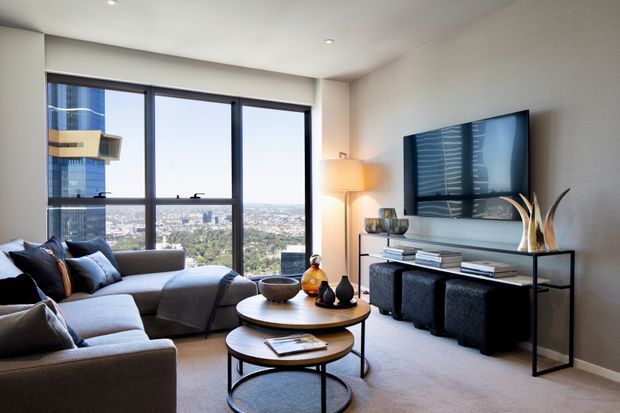 A contemporary high-rise living room with floor-to-ceiling windows overlooking a dense urban landscape. The room features a large sectional sofa, nested round coffee tables, a wall-mounted television, a slim black console, textured light walls, recessed ceiling lights, and a floor lamp. The interior is neutral, refined, and warmly lit, with a strong connection to the city view.