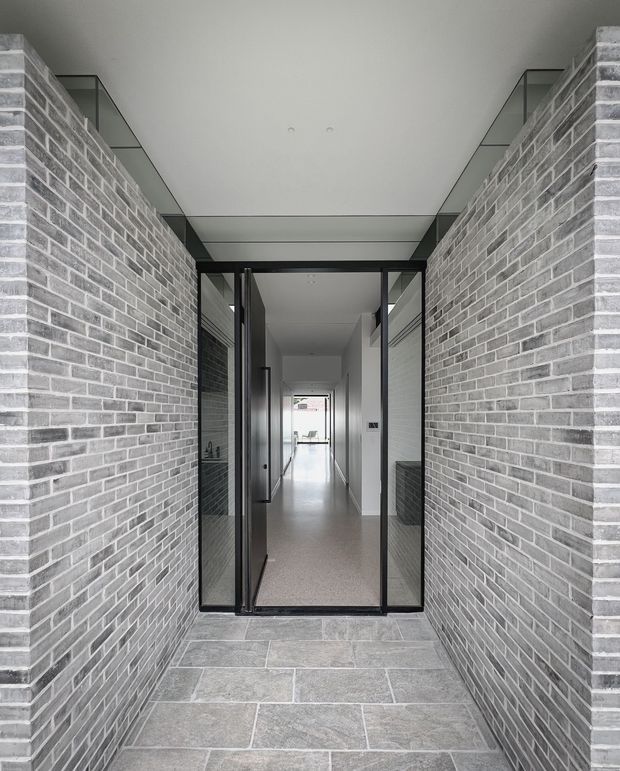 A minimalist residential entryway viewed from an exterior covered approach. Grey brick walls frame a glazed black-framed entrance, with large-format grey stone paving leading into a long, bright interior corridor. The composition emphasizes strong linear perspective, restrained materials, and abundant glazing.