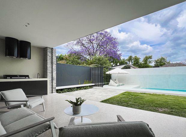 A covered contemporary outdoor living area opens onto a landscaped courtyard with a rectangular swimming pool. The space includes built-in dark cabinetry and an outdoor kitchen or barbecue zone, pale textured flooring, lounge seating, a round coffee table, and a large white retractable-style canopy over the poolside area. A dark vertical metal privacy screen and exposed light brick column frame the view. The palette is predominantly light grey, white, charcoal, and green, with a jacaranda tree visible beyond the property.