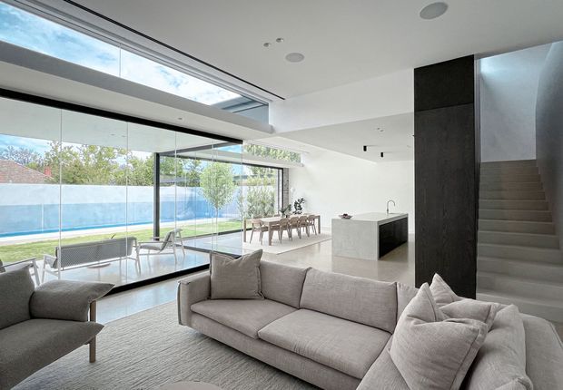 A bright, spacious contemporary open-plan living area with a large neutral sectional sofa and armchair in the foreground. Full-height glazing opens toward a pool and garden, while a dining area and minimalist kitchen island sit beyond. A dramatic rooflight and clerestory glazing bring in abundant daylight, and a dark vertical feature wall frames the staircase.