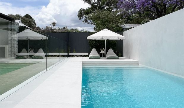 A contemporary outdoor swimming pool courtyard with a long rectangular turquoise pool, pale concrete paving, frameless glass balustrade, white rendered boundary walls, and a shaded lounge area with white umbrellas and sculptural outdoor seating. Dark fencing, mature trees, and a jacaranda add contrast around the private pool setting.