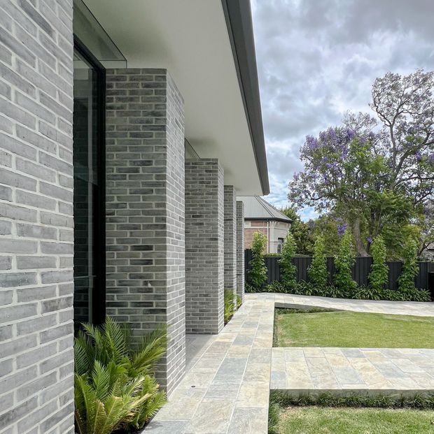 Contemporary residential exterior showing a long grey brick facade with deep recessed floor-to-ceiling glazing, projecting roof eaves, and a paved walkway leading through a landscaped courtyard. Large-format pale stone paving, clipped lawn, black fencing, established planting, and neighbouring homes are visible under an overcast sky.