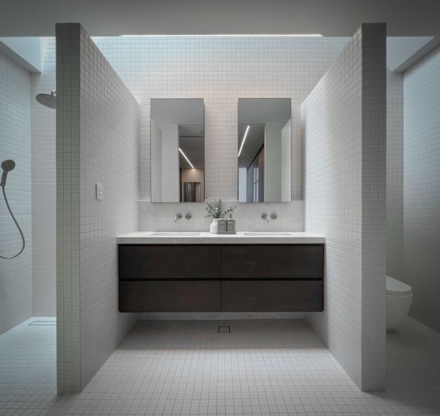 A contemporary bathroom with a floating dark timber vanity, pale stone benchtop, twin rectangular mirrors, wall-mounted taps and basin fixtures, and extensive small-format white tiled walls and floor. Open shower areas are visible on the left and a toilet is partially visible on the right. A linear skylight or clerestory above the tiled central volume provides soft natural light.
